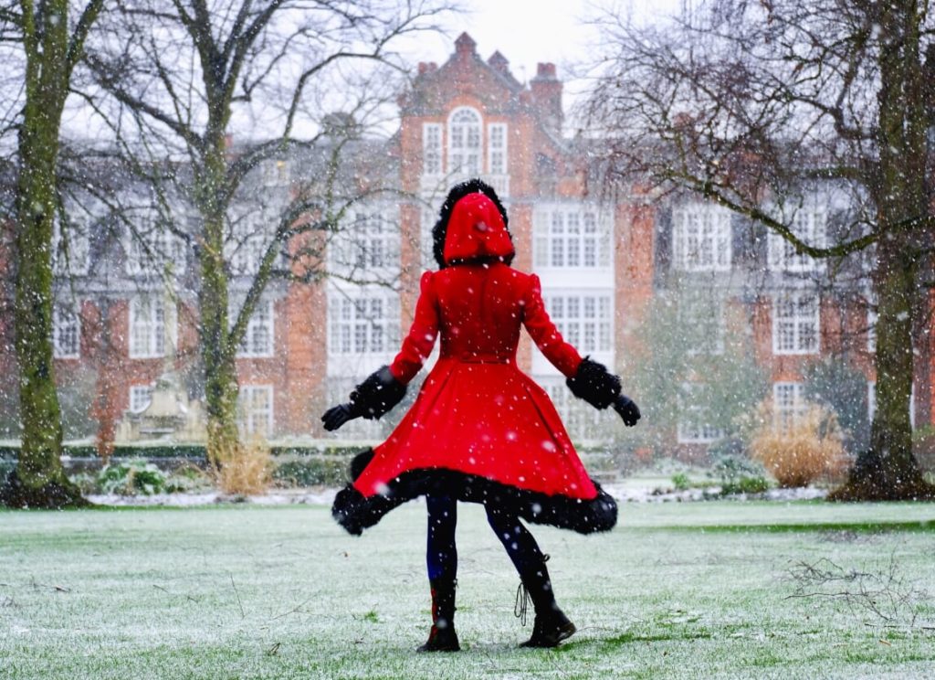 A person in a red coat, fringed with black faux-fur is in the middle of a twirl on a snowy, grassy lawn. The building of Peile is visible in the background through snowflakes falling. 
