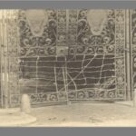 Ornate bronze gates with the lower panels missing and bent and the gap filled with barbed wire