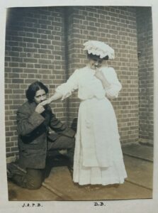 A scan of a black and white photograph. A woman in Edwardian clothes stands, one finger to her lips, her hand outstretched behind her. A dark-haired person in a tweed jacket and trousers and a fake moustache is on one knee behind her, kissing her hand. Both are looking at the camera. They appear to be standing on a lead roof, with a brick wall behind them.
